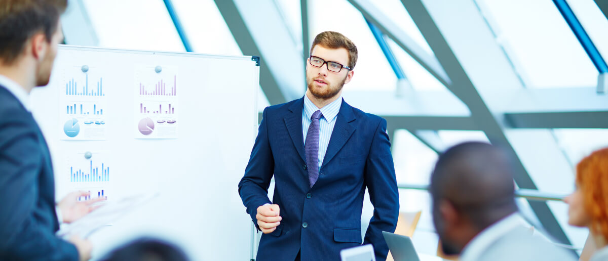 a business professional giving a presentation in front of a whiteboard with various charts and graphs, to a group of attentive colleagues